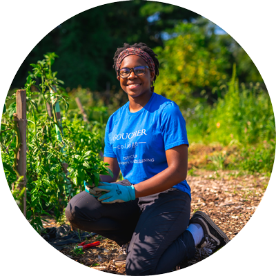 A Goucher students kneels among vegetable plants at a community garden project.